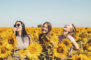 Three women laughing and smiling in a field of sunflowers on a sunny day while wearing Nöz colorful SPF sunscreen on their noses 